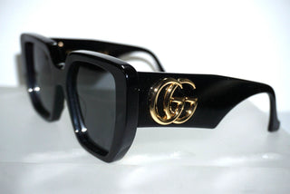 Black square sunglasses with a gold interlocking GG logo on the temple, displayed on a reflective white surface.