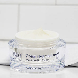 A clear cosmetic jar of Obagi Hydrate Luxe Moisture-Rich Cream with white cream visible on top, set against a clean white tiled background.